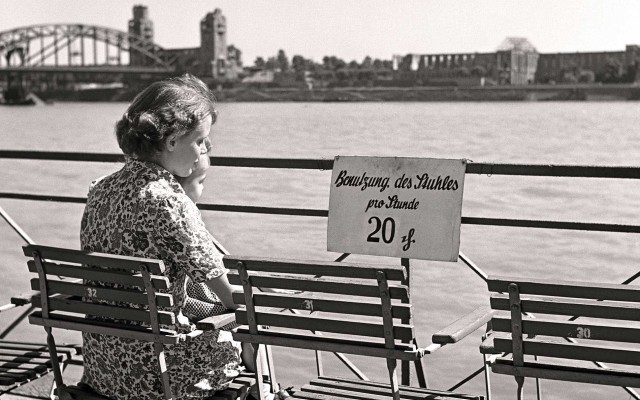 Frau sitzt mit einem Kind auf dem Schoß am Kölner Rheinufer mit Blick auf die Hohenzollernbrücke. Am Geländer ein Schild "Benutzung des Stuhls pro Stunde 20 Pfennig"