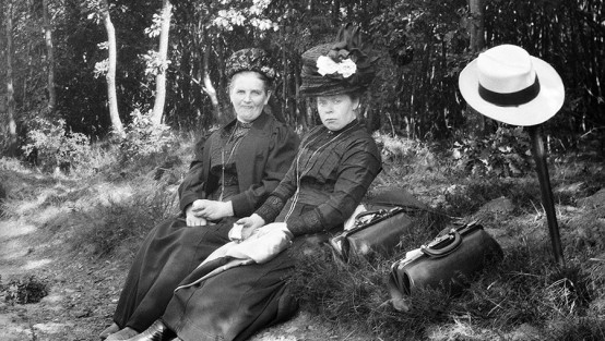 Zwei Frauen in historischer Bekleidung und mit Hüten sitzen während einer Wanderung am Wegrand und ruhen sich aus