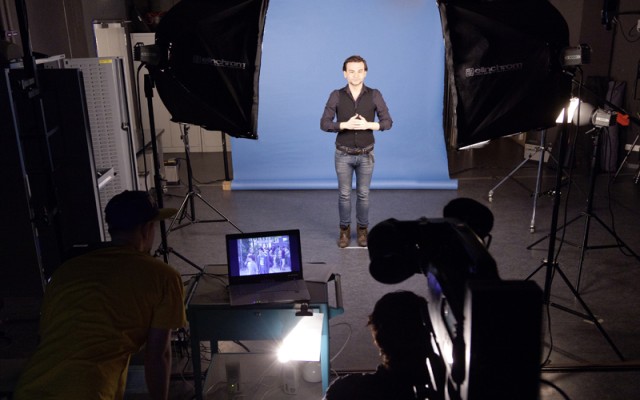 Ein Gebärdensprachdolmetscher vor einer Kamera im Fotostudio. Hinter ihm eine blaue Wand. Fotostudio mit Setaufbau für den Dreh von Gebärdensprachvideos.