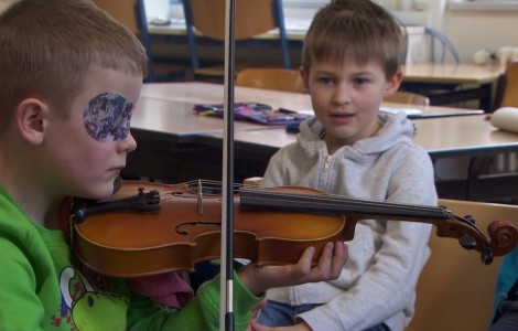 Kleiner Junge spielt auf einer Geige. Ein anderer schaut ihm zu.