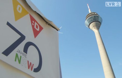 Links ein Banner mit der Schrift 70 Jahre NRW aus der Untersicht, rechts der Düsseldorfer Fernsehturm.